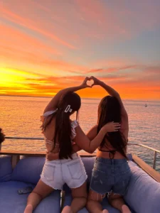 Two Women forming a heart shape with their hands while sitting on the bow of a Lover Lips Yachts boat during a colorful sunset in La Pa, Baja California Sur