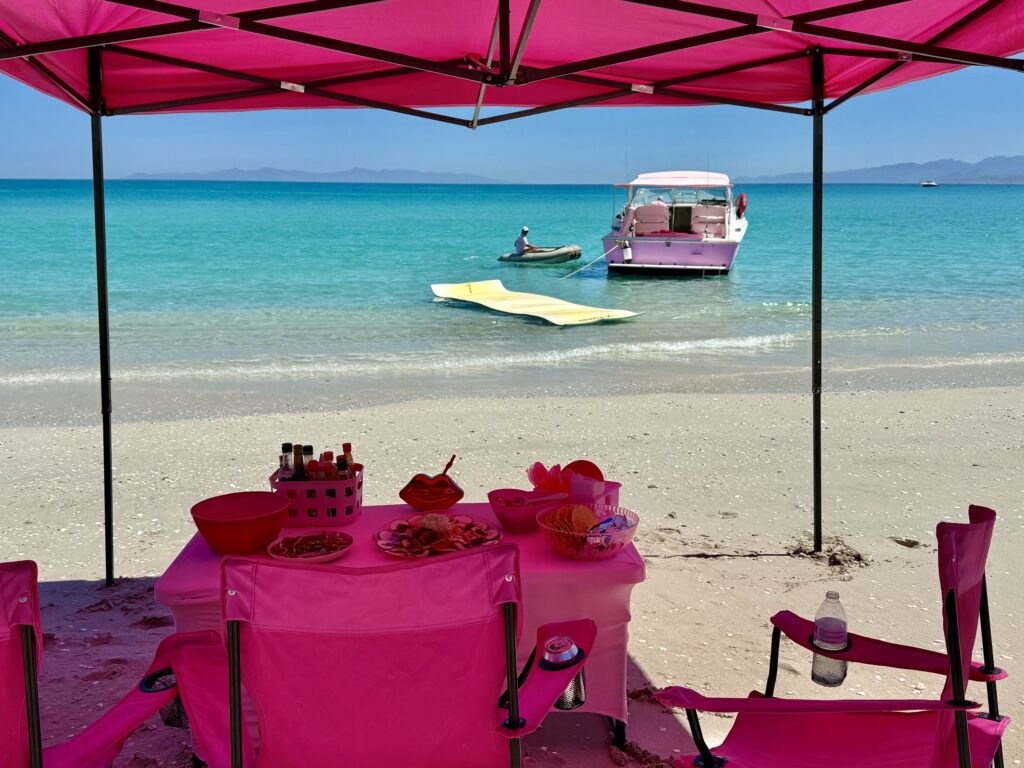 Private Beach Setup at Balandra Beach | Lover Lips Yachts La Paz Mexico - Lover Lips Yachts Pink beach party setup under a canopy with a table of snacks and drinks, turquoise sea in the background, a pink boat nearby.