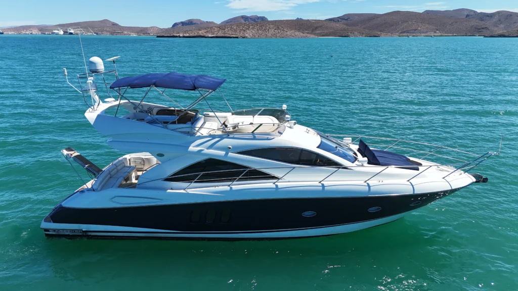 White luxury motor yacht with blue canopy on calm turquoise water, coastal hills in the background.