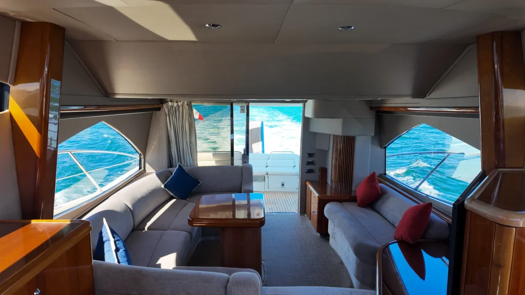 Interior view of a luxury yacht lounge with gray upholstery, wooden tables, and red and blue cushions, facing large windows to the sea.